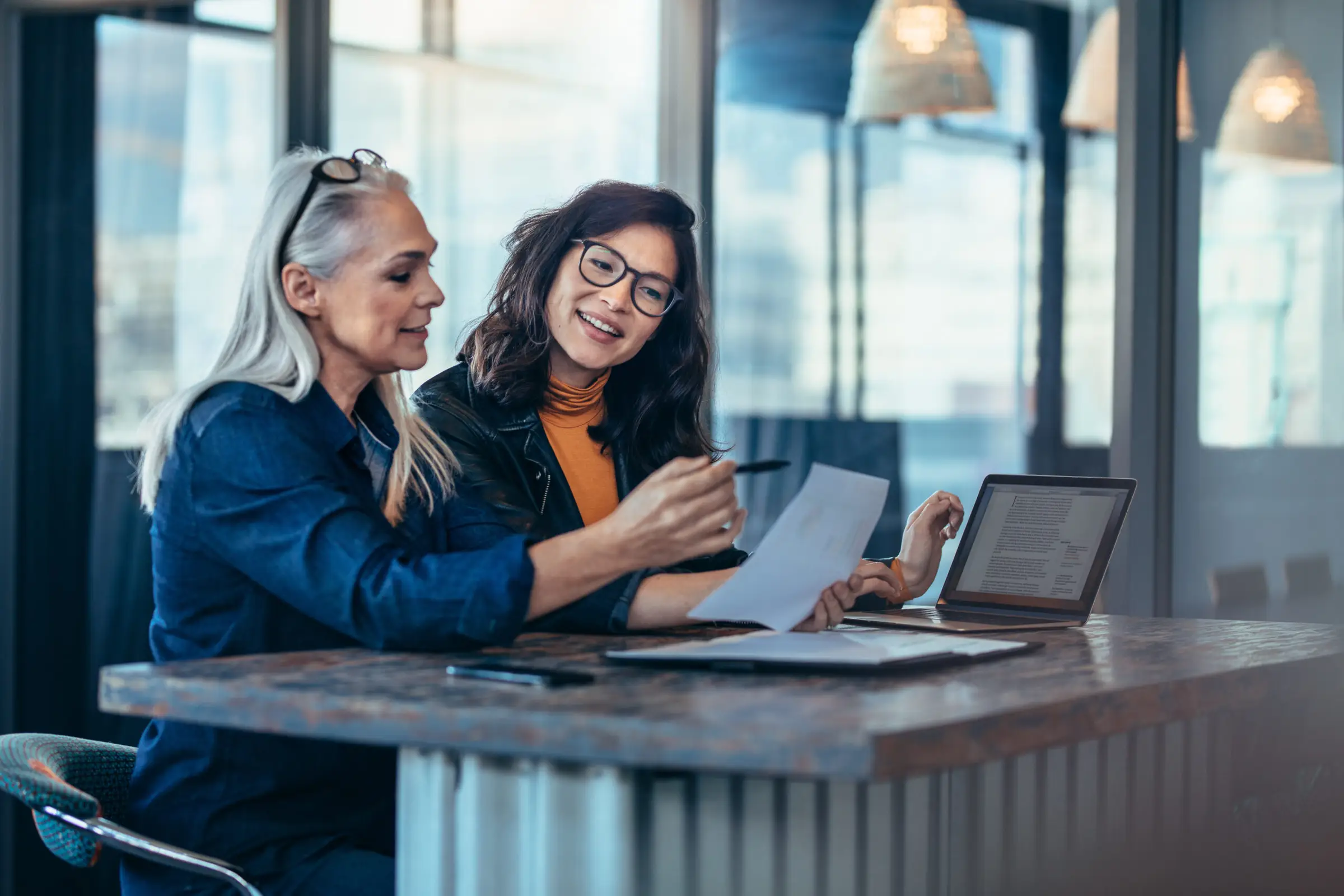 Two women from Light Side Wellness Co. sit at a table in an office, reviewing a document together, focused on enhancing mental health programs. One woman holds a pen, and an open laptop is beside them. Large windows in the background let natural light flood the room.