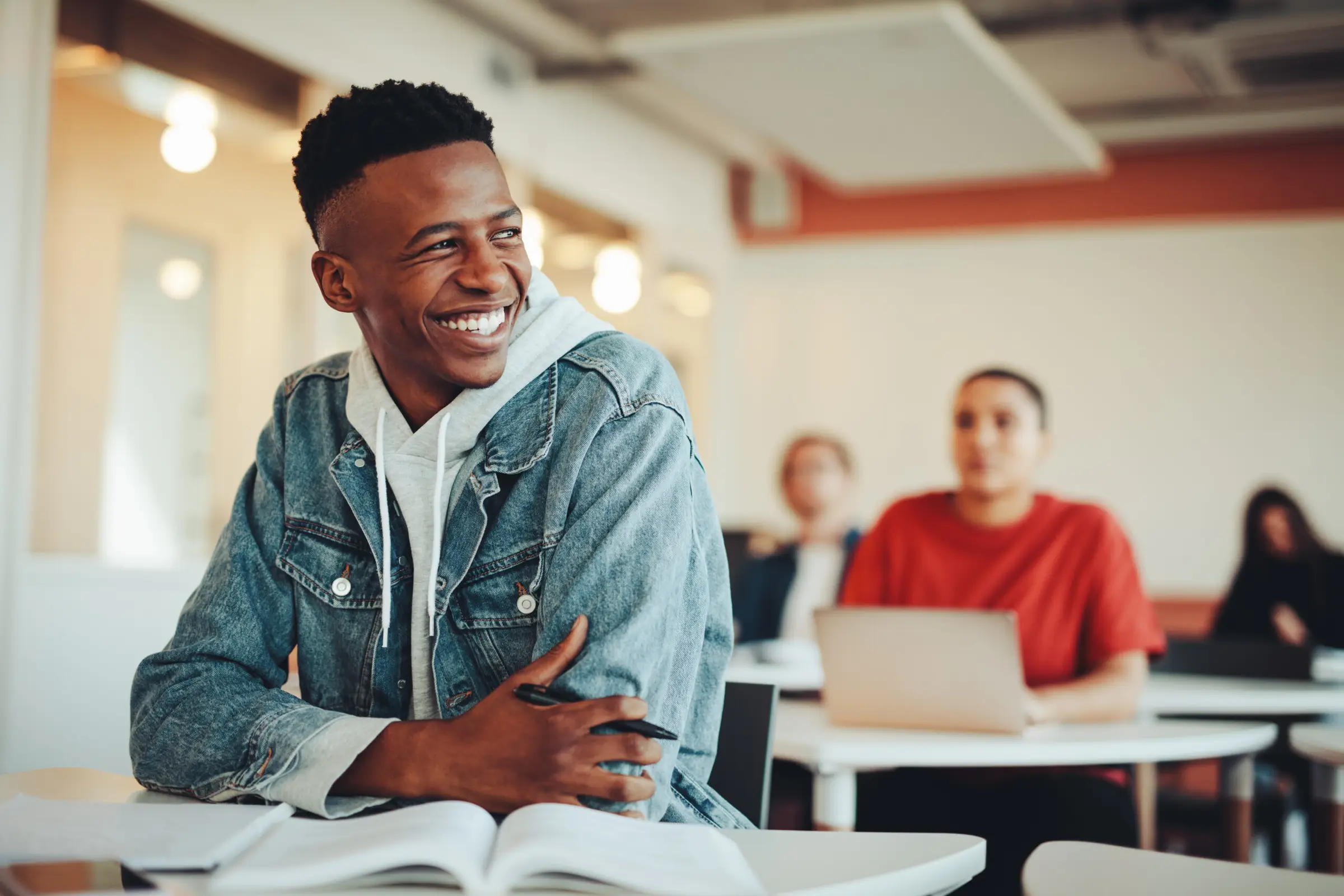 A person in a denim jacket smiles while sitting at a desk in a classroom at Light Side Wellness Co. An open book and pen are in front of them. In the background, other students are seated, one with a laptop. The room is well-lit and modern.