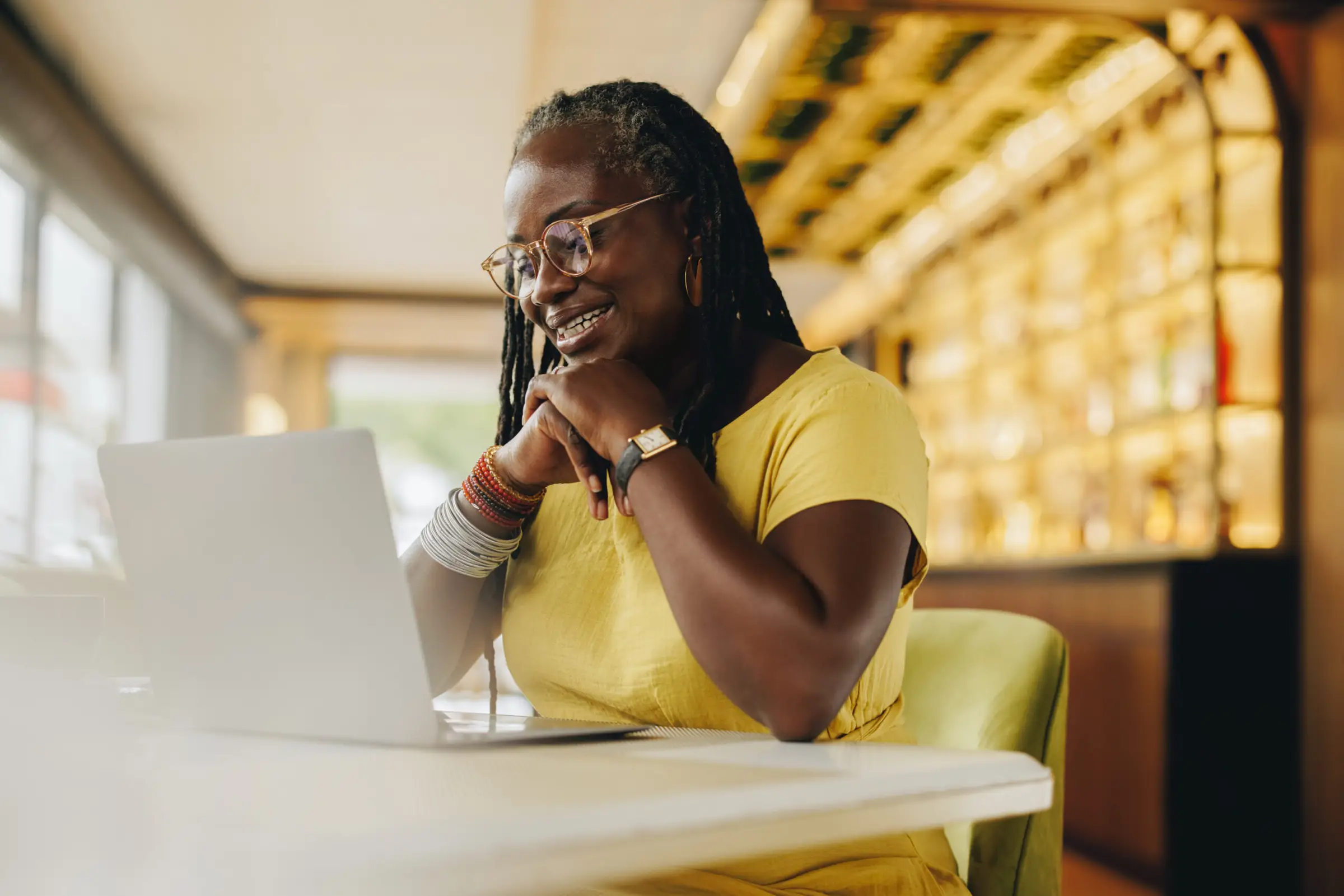 A woman with long hair and glasses sits at a table, smiling and looking at a laptop. She's wearing a yellow top, resting her chin on her hands. The well-lit room with shelves suggests a calming space, perfect for focusing on mental health and exploring Portland psychiatry resources.