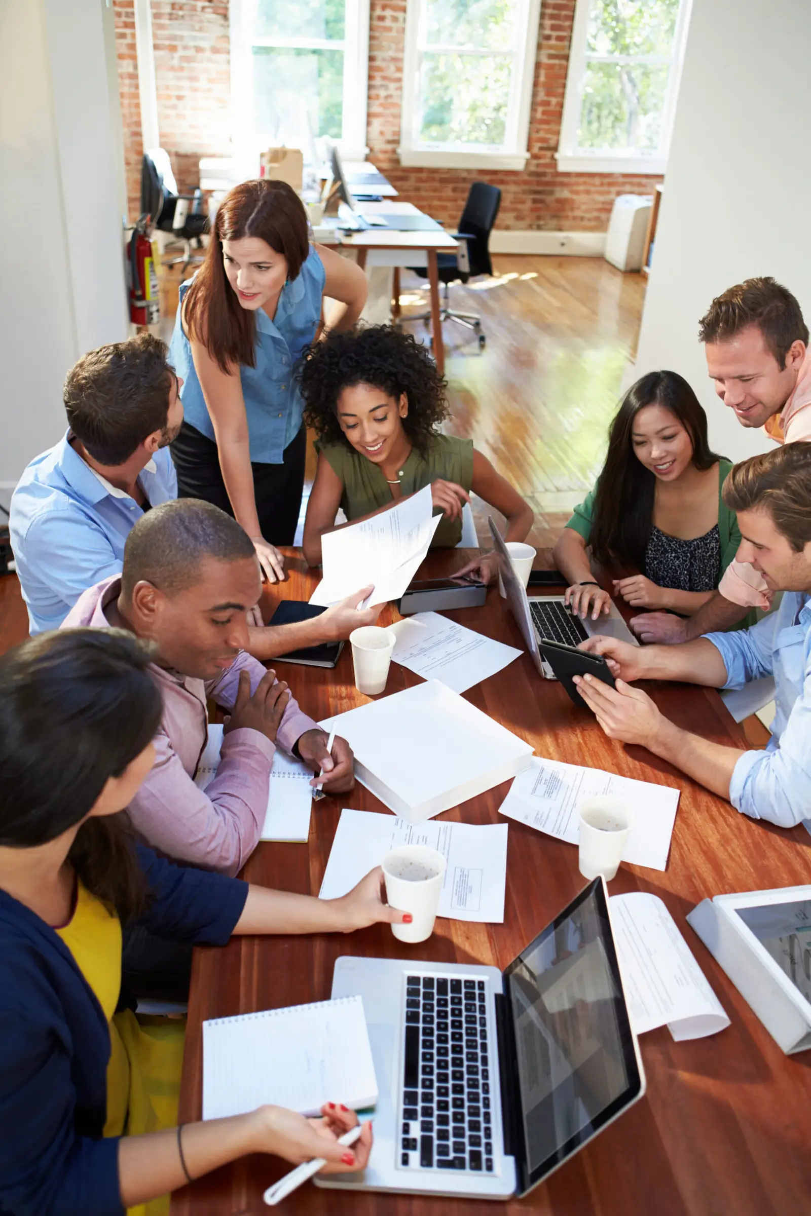 A diverse group of people is gathered around a table in a modern office, working collaboratively on mental health initiatives. They have laptops, papers, and coffee cups in front of them, engaged in discussion and planning. The room is bright with natural light.