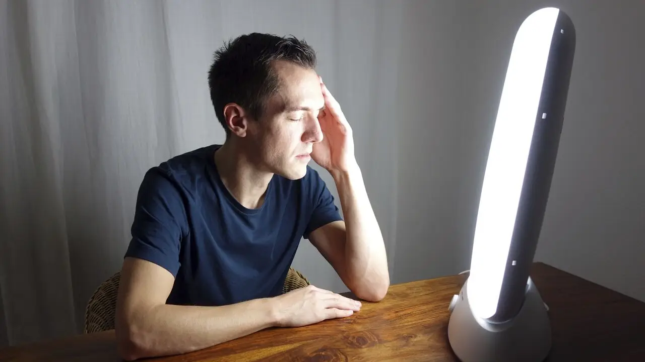 A person with short dark hair sits at a wooden table, eyes closed, facing a bright Light Side Wellness Co therapy lamp. They rest their left hand on their forehead, wearing a dark t-shirt. The background is a plain white curtain.