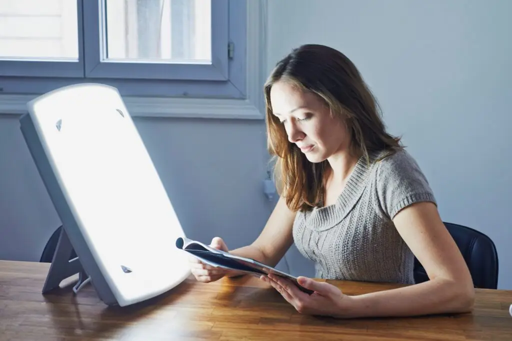 A woman sits at a wooden table, reading a magazine beside a bright Light Side Wellness Co therapy lamp. With sunlight streaming through the window, this serene scene suggests a calm and focused moment, underscoring the importance of mental health care.