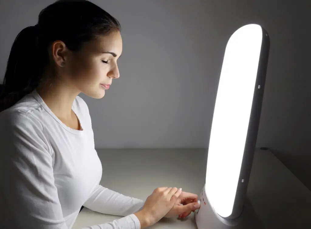 A woman in a white shirt sits with her eyes closed, facing a bright light therapy lamp on a table. The dimly lit surroundings highlight the lamp's glow, creating a calming ambiance that supports mental health and alleviates symptoms often discussed in Portland psychiatry sessions.