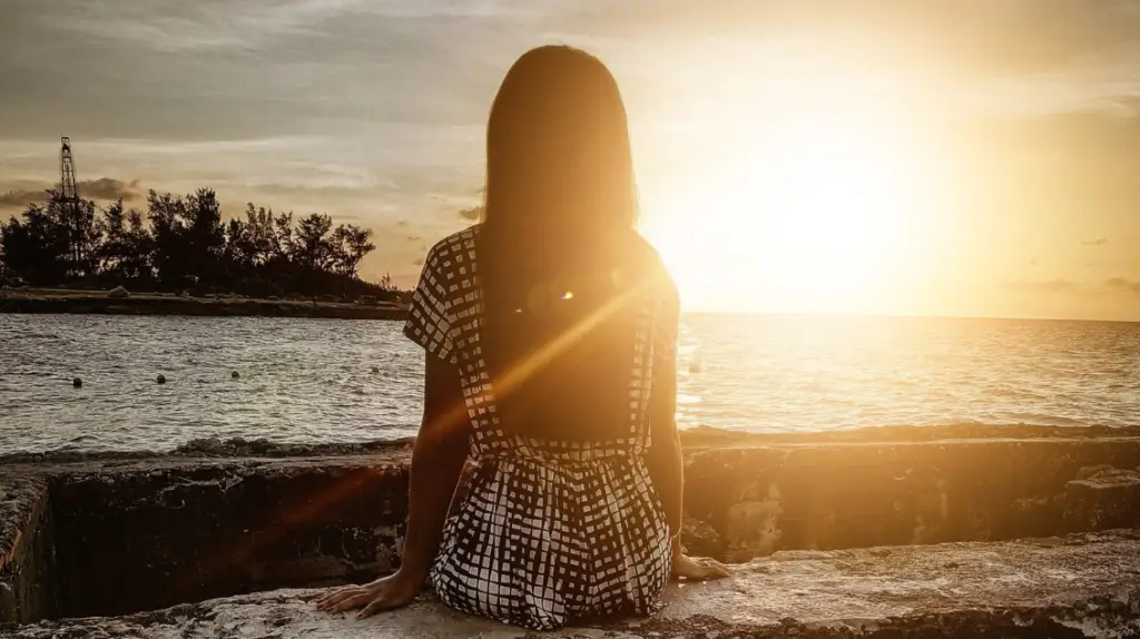 A person with long hair sits on a stone wall, facing the calm ocean. The sun sets on the horizon, casting a warm glow reminiscent of Light Side Wellness Co's serene approach to mental health. Trees and a tower are silhouetted in the distance, exuding tranquility and reflection.