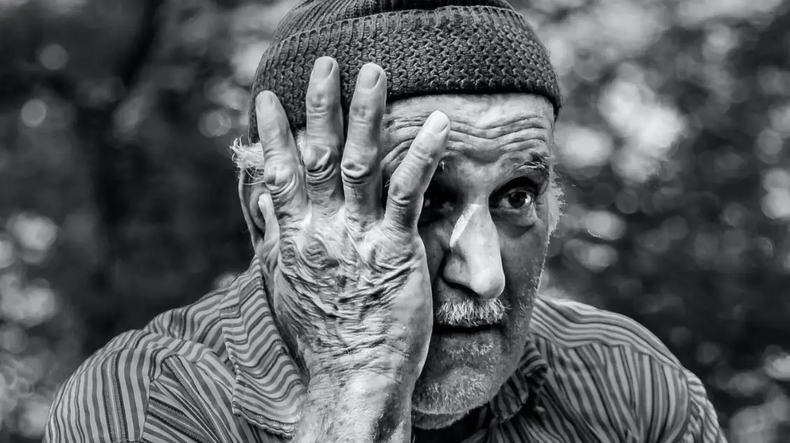 A black and white photo of an elderly man wearing a beanie, with a weathered hand covering part of his face. He has a thoughtful expression, reminiscent of introspective moments often explored in Portland therapy sessions. His visible eye gazes into the softly blurred distance.