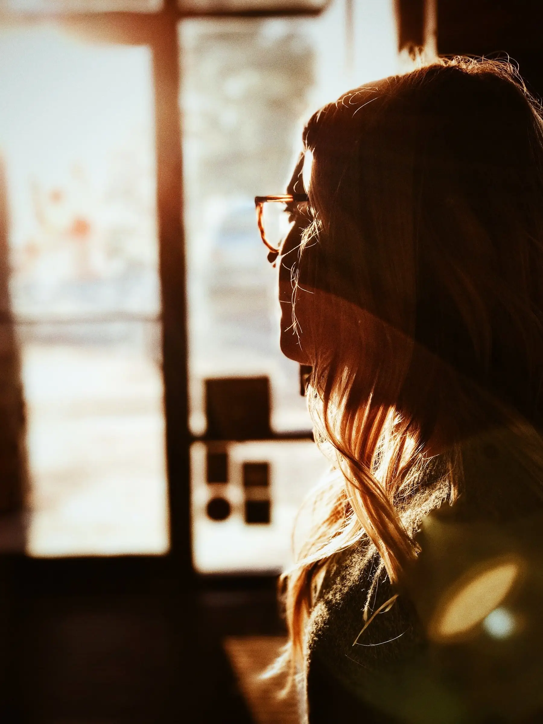 A silhouette of a woman with long hair and glasses stands in front of a sunlit window, embodying the tranquility often sought in Portland therapy. The warm glow creates a peaceful atmosphere, casting gentle shadows and highlights around her profile.